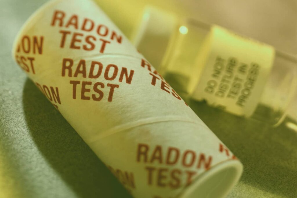 A close-up of a cylindrical radon test kit with "RADON TEST" printed repeatedly across its surface, positioned next to a blurred yellow warning label that reads "DO NOT DISTURB TEST IN PROGRESS."
