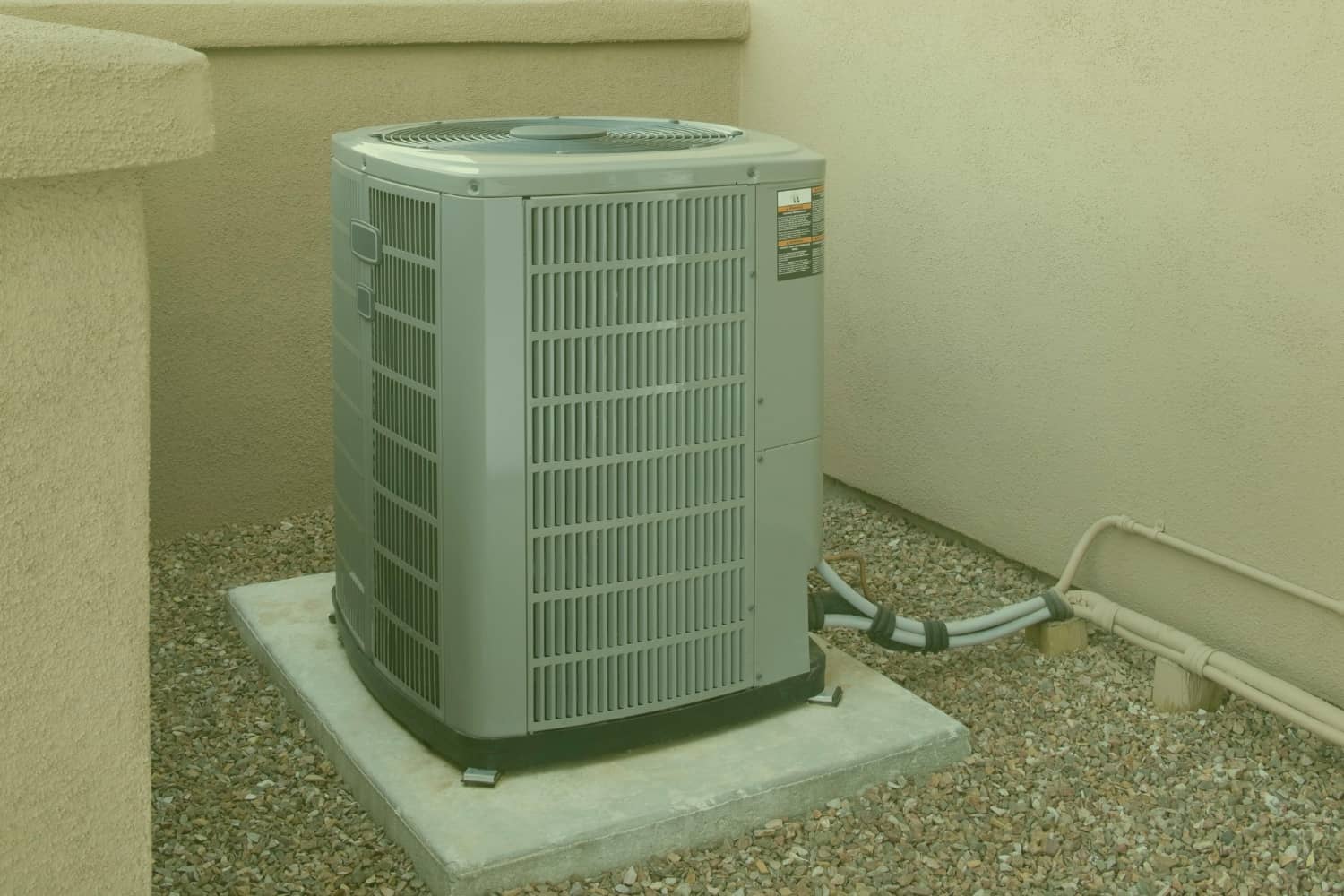 A gray outdoor central air conditioner condenser unit sits on a concrete pad surrounded by gravel next to a beige stucco exterior wall.