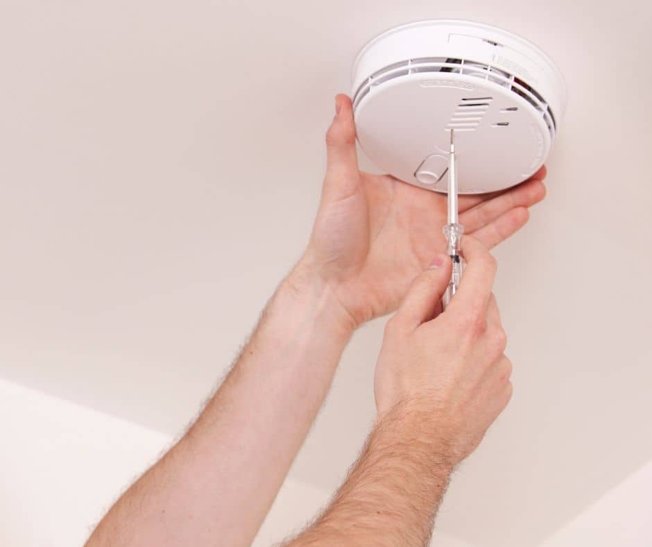 Hands using a small screwdriver to install or service a ceiling-mounted smoke detector